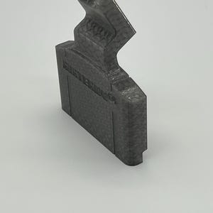 Puede incluir: Un objeto de plástico gris oscuro impreso en 3D con forma de cartucho y superficie texturizada. La cara frontal presenta el texto en relieve NINTENDO 64. La sección superior tiene un diseño de corte angular y forma rectangular.