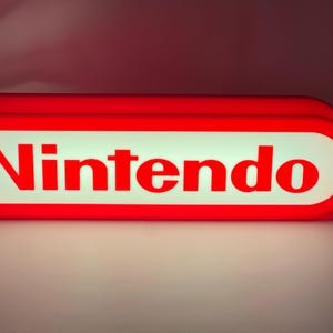 Może przedstawiać: Czerwono-biały znak Nintendo. Znak jest prostokątny z zaokrąglonymi krawędziami. Słowo "Nintendo" jest napisane czerwonymi, pogrubionymi literami na białym tle. Znak jest podświetlony, z jasną poświatą.