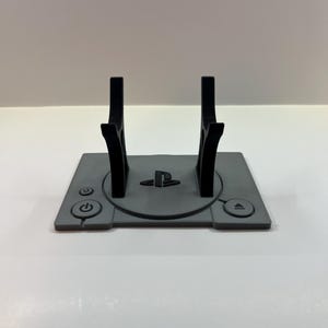 May include: A gray and black PlayStation console stand. The stand features a gray base with a power button, eject button, and the PlayStation logo. Two black vertical supports rise from the base to hold the console.