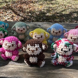 Puede incluir: Una colección de muñecos de mono de ganchillo coloridos en varios tonos de azul, verde, morado, rosa, amarillo, marrón y rojo. Cada mono tiene una cara blanca y está sentado sobre una superficie de madera. Los monos hechos a mano tienen un diseño lindo y caprichoso.