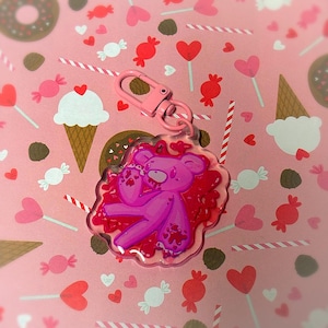 May include: A pink acrylic keyring with a cartoon bear design. The bear is surrounded by a red, heart-shaped design and has a pink clasp. The background is a pink pattern with sweets and hearts.