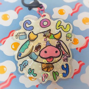 May include: A clear, cow-shaped keychain with the words "Cow Furry" in colorful bubble letters. The cow has a pink nose and tongue, with a small tag that says "69". The background is a light blue with images of fried eggs and bacon.