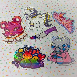 May include: A selection of vibrant stickers with diverse designs. The stickers include a pink tiger, a white and purple pony, a knife, a sea slug, a rainbow creature, and a bear with a transgender flag. The stickers have a holographic finish.