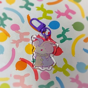 Puede incluir: Un llavero acrílico transparente con un personaje de ratón de dibujos animados con un atuendo lavanda y un cierre morado. El ratón tiene ojos rosados y una cara blanca. El fondo es un patrón colorido de animales de globos.