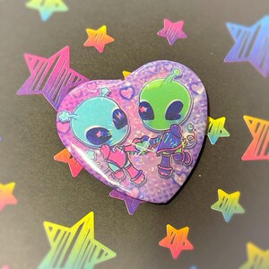 Può includere: Un bottone a forma di cuore con due alieni dei cartoni animati che si tengono per mano. Uno è blu e rosa, l'altro verde e viola. Il bottone ha un effetto olografico glitterato. Lo sfondo è nero con adesivi a forma di stella colorati.