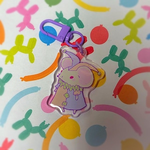 Puede incluir: Un llavero acrílico transparente con un ratón de dibujos animados vestido de bufón. El ratón tiene orejas y nariz rosas, vistiendo un atuendo multicolor. El llavero tiene un cierre morado y un pequeño anillo. El fondo tiene un patrón de globos coloridos.