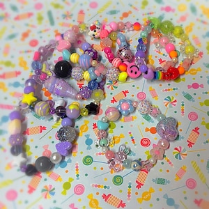 May include: A collection of colorful beaded bracelets and necklaces. The jewelry features a variety of beads in pastel colors, including pink, purple, blue, and yellow. Some beads are shaped like stars, flowers, and ice cream cones. The background is a light blue pattern with candy.