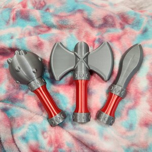 May include: Three toy weapons: a mace, a double-bladed axe, and a bladed weapon. The weapons are gray with red handles. The mace has a ball head with protruding studs. The axe has a cross-shaped handle. The bladed weapon has a curved blade.