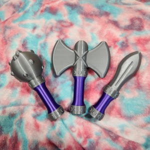 May include: Three toy weapons: a mace, a double-bladed axe, and a spear. The weapons are gray with purple handles and silver accents. The objects are arranged on a colorful, tie-dye patterned surface.
