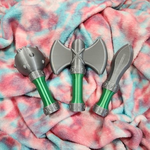May include: Three toy weapons: a mace, a double-bladed axe, and a spear. The weapons are gray with green handles and decorative silver accents. They are arranged on a pink, blue, and white tie-dye patterned blanket.