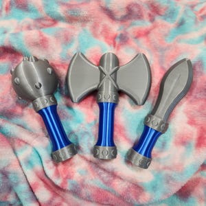 May include: Three toy weapons: a mace, a double-bladed axe, and a spear. The weapons are gray with blue handles and silver accents. The mace has a spiked head. The axe has a cross-shaped handle. The spear has a pointed blade.