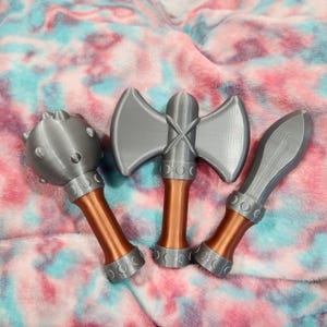May include: Three toy weapons: a mace, a double-bladed axe, and a dagger. The weapons are gray with copper-colored handles. The mace has a spiked ball head. The axe has a cross-shaped handle. The dagger has a curved blade.