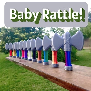 May include: A row of colorful baby rattles shaped like axes. Each rattle has a gray axe head and a handle with a different color, including red, blue, green, and yellow. The text "Baby Rattle!" is above the rattles.