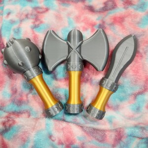 May include: Three toy weapons: a mace, a double-bladed axe, and a spear. The mace has a gray head with rounded studs and a gold handle. The axe and spear have gray blades and gold handles. All three have a decorative silver band.