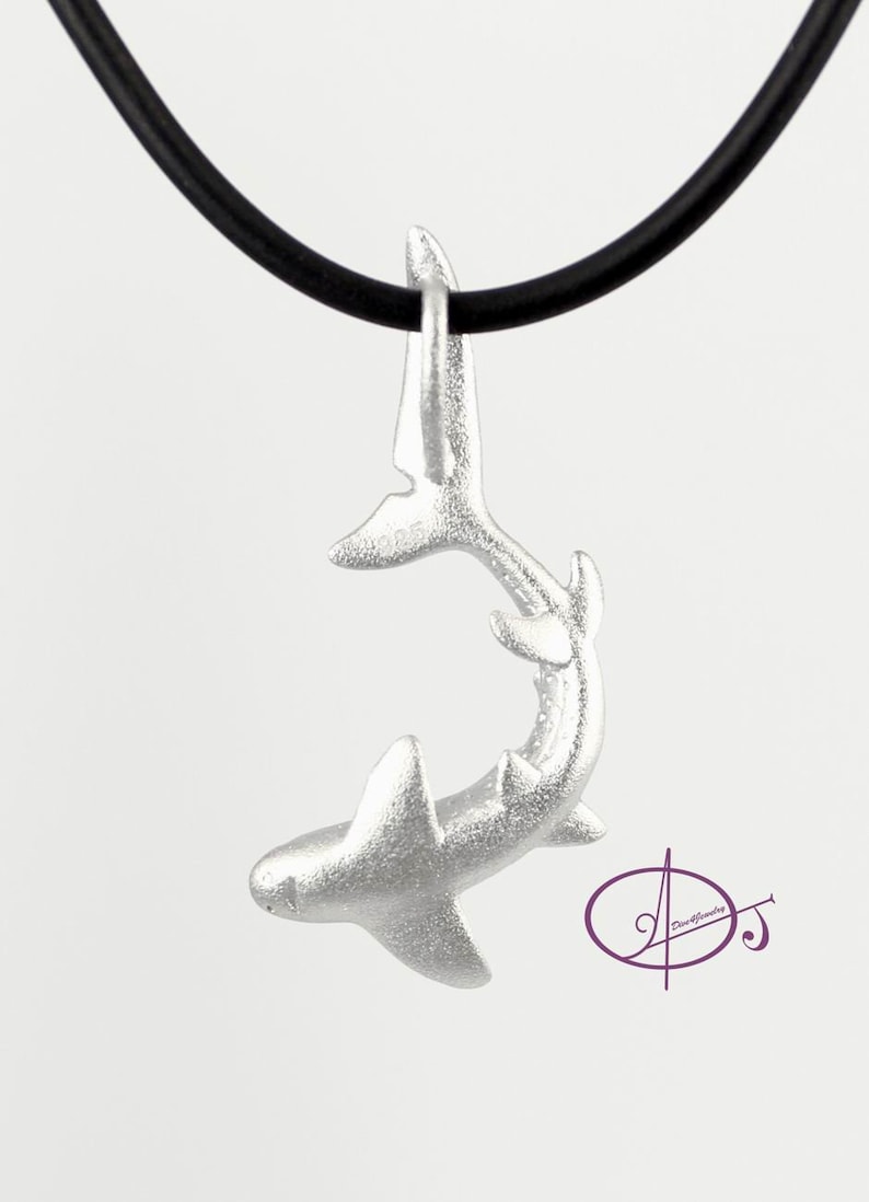 May include: A silver shark pendant necklace on a black cord. The shark is in a curled position with its tail up and its head down. The pendant is detailed and realistic.