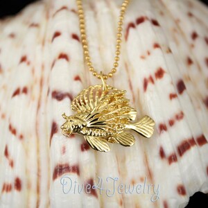 K&ouml;nnte beinhalten: Goldene  L&ouml;wenfisch-Anh&auml;nger-Halskette an einer goldenen Kette. Der Anh&auml;nger ist detailliert und filigran gearbeitet, mit einem realistischen Design. Die Halskette wird auf einer wei&szlig;en Muschel mit braunen Markierungen pr&auml;sentiert.