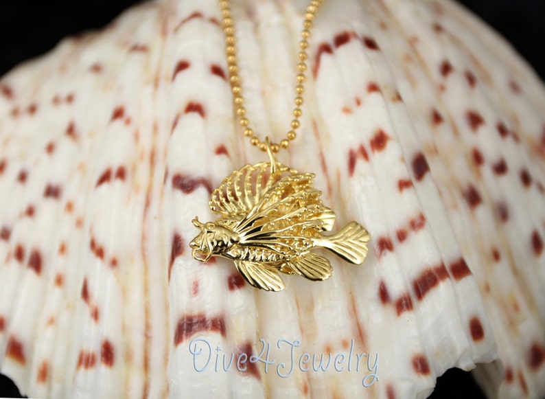 K&ouml;nnte beinhalten: Goldene  L&ouml;wenfisch-Anh&auml;nger-Halskette an einer goldenen Kette. Der Anh&auml;nger ist detailliert und filigran gearbeitet, mit einem realistischen Design. Die Halskette wird auf einer wei&szlig;en Muschel mit braunen Markierungen pr&auml;sentiert.