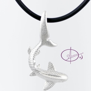 May include: A silver shark pendant necklace with a black cord. The shark is in a curled position with its tail up and its head down. The pendant is detailed with a textured surface.