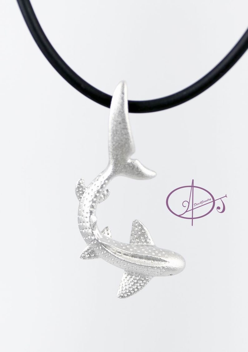 May include: A silver shark pendant necklace with a black cord. The shark is in a curled position with its tail up and its head down. The pendant is detailed with a textured surface.