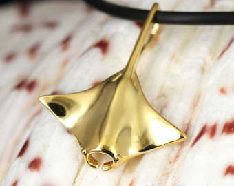 May include: A gold-colored pendant in the shape of a manta ray, suspended from a black cord. The pendant has a smooth, reflective surface and is set against a textured, off-white background. The jewelry is a marine-themed accessory.