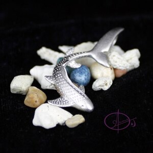 May include: A silver whale shark figurine with a spotted pattern, posed in a swimming position. The figurine is surrounded by small white, brown, and blue stones.