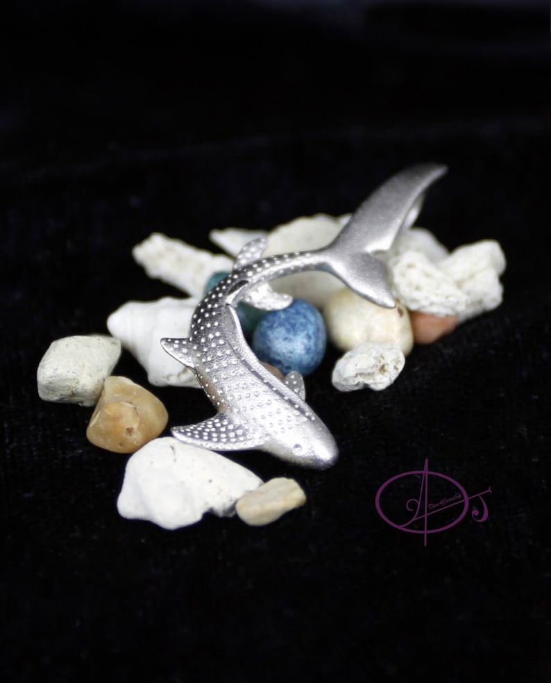 May include: A silver whale shark figurine with a spotted pattern, posed in a swimming position. The figurine is surrounded by small white, brown, and blue stones.
