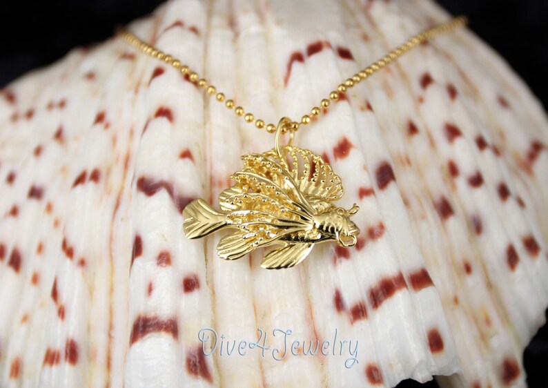 K&ouml;nnte beinhalten: Goldene  L&ouml;wenfisch-Anh&auml;nger-Halskette auf einem wei&szlig;en Muschelhintergrund. Der Anh&auml;nger ist detailliert und filigran, mit einer strukturierten Oberfl&auml;che. Die Halskette besteht aus einer zarten Kette.