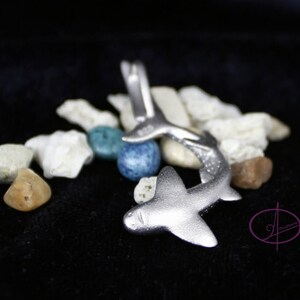 May include: A silver shark charm pendant with a detailed design. The charm is surrounded by small white and blue stones.