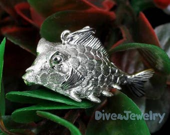 May include: Silver fish charm pendant with a detailed, textured design. The charm is shaped like a fish with a slightly open mouth and a fin on its back. The charm is set against a green leafy background.