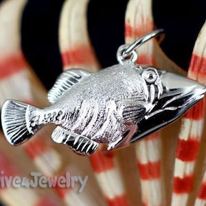 May include: A silver fish pendant charm with a detailed, textured design. The pendant is shaped like a triggerfish and has a loop at the top for attaching to a chain or bracelet.