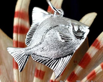 May include: A silver-colored fish pendant with detailed fins and scales. The pendant is suspended from a small loop and is set against a backdrop of cream and red striped elements. The pendant is likely jewelry.