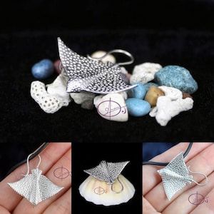 Puede incluir: Collar con colgante de raya en plata, con superficie texturizada. El colgante se muestra sobre un fondo negro con pequeñas piedras blancas y azules.