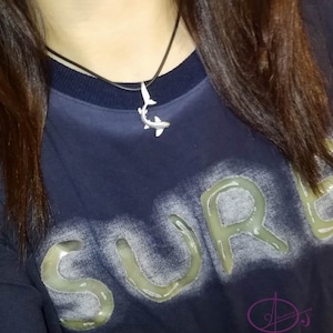 May include: A close-up of a person wearing a navy blue shirt with the word "SURF" printed in white on the front. The person is wearing a silver shark pendant necklace.