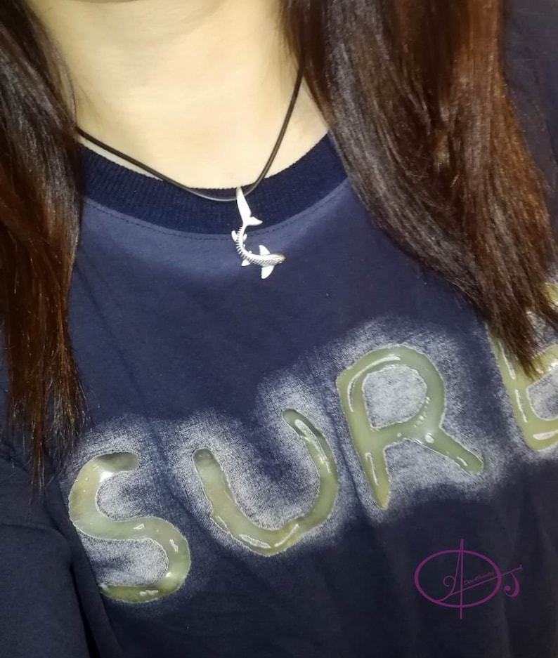May include: A close-up of a person wearing a navy blue shirt with the word "SURF" printed in white on the front. The person is wearing a silver shark pendant necklace.