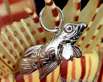 May include: Silver charm in the shape of a fish with intricate details. The charm is hanging from a silver loop and is set against a background of orange and white stripes.