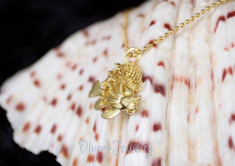 K&ouml;nnte beinhalten: Eine goldene  L&ouml;wenfisch-Anh&auml;nger-Kette an einer goldenen Kette. Der Anh&auml;nger ist detailliert und realistisch, mit Flossen und Schuppen. Die Kette wird auf einer wei&szlig;en Muschel mit braunen Markierungen gezeigt.