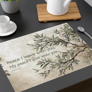 May include: A rectangular placemat featuring a beige backdrop and a watercolour olive branch design. The text "Peace I leave with you, My peace I give unto you. JOHN 14:27" is printed on the placemat. A white teacup, saucer, and teapot are also visible.