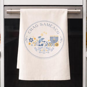 Könnte beinhalten: Cremefarbenes Küchentuch mit dem Schriftzug "CHAG SAMEACH" in einem kreisförmigen Design. Das Design umfasst einen Davidstern, einen Vogel, eine Menora und florale Elemente in Blau und Gelb. Das Tuch hängt an einem Ofengriff.