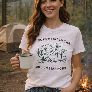 Może przedstawiać: Biała koszulka z zieloną grafiką przedstawiającą krajobraz górski i napisem "SUKKOTIN' IN THE BILLION STAR HOTEL". Kobieta trzyma kubek przed ogniskiem i namiotem.