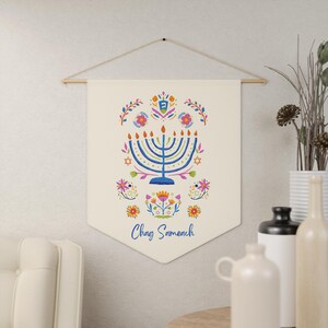 Puede incluir: Colgante de pared color crema con un diseño floral y una menorá azul. El diseño incluye flores naranjas, rosas y azules, y el texto "Chag Sameach" en escritura azul. El colgante está suspendido de una barra de madera.