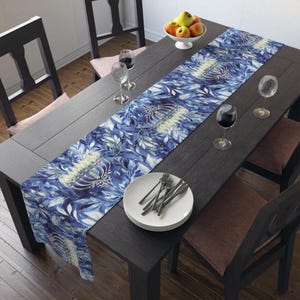 May include: A blue and white floral table runner with a gold design, placed on a dark wooden table. The table is set with plates, silverware, and wine glasses. A bowl of fruit sits at the top.
