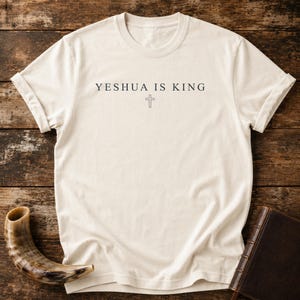 Puede incluir: Camiseta color crema con el texto "YESHUA IS KING" y una cruz. Las mangas están dobladas. Un libro marrón y un cuerno son visibles sobre una superficie de madera en el fondo.