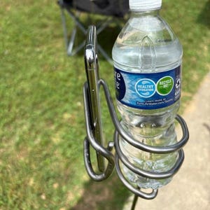 Könnte beinhalten: Eine transparente Plastikwasserflasche mit weißem Deckel wird in einem silbernen Metallhalter gehalten. Ein Smartphone wird ebenfalls in der Halterung gehalten. Der Halter ist an einer Stange befestigt, mit einem Klappstuhl und anderen Getränken im Hintergrund.