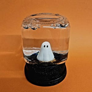 Puede incluir: Un globo de nieve con temática de Halloween con una pequeña figura de fantasma blanco dentro de un frasco de vidrio lleno de agua. El frasco se asienta sobre una base negra contra un fondo naranja. El fantasma tiene dos ojos negros.