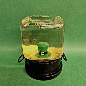 Può includere: Un contenitore di vetro trasparente riempito di liquido giallo e un cappello da folletto verde. Il contenitore è posto su un supporto nero e rotondo con un manico. Lo sfondo è verde.