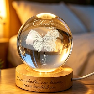 May include: A clear glass sphere night light resting on a circular wooden base. The sphere features an etched butterfly and floral design with the text "Chapter THIRTY" and "Victoria". The wooden base is engraved with "New chapter, new dreams, same beautiful you".