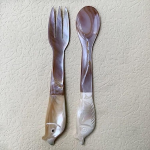 May include: A fork and spoon set with a fish-shaped handle. The utensils have a pearlescent finish with brown and white tones. The fork has three tines, and the spoon has a rounded bowl. The handles are detailed with fish features.