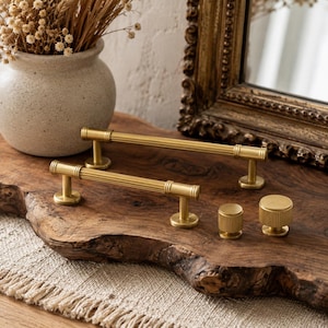 Peut inclure: Deux poignées de placard en métal doré avec un design cylindrique nervuré et deux boutons de placard ronds et cannelés assortis. La quincaillerie présente une finition métallique brossée et une texture linéaire sur les poignées et les boutons.