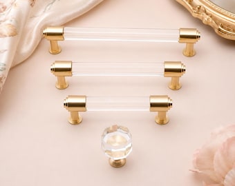 May include: Three clear acrylic cabinet pulls with brushed gold metal ends in varying lengths, arranged horizontally, and one matching clear acrylic faceted cabinet knob with a brushed gold base, all featuring a cylindrical design.