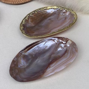 以下が含まれることがあります： 2つの楕円形の装飾皿。1つは茶色と紫の色合いの真珠光沢のある表面を持っています。もう1つは金色の装飾的な縁があります。これらの皿はおそらく真珠母貝で作られています。